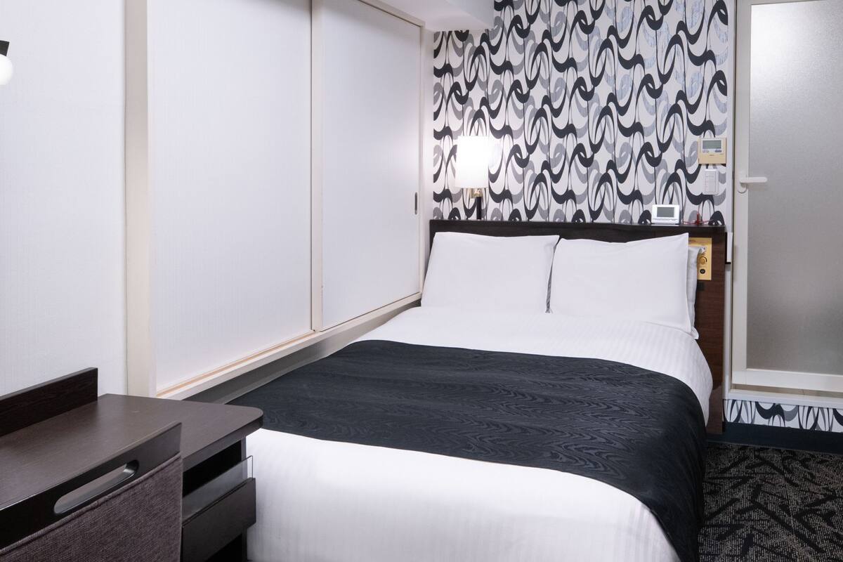A hotel room with a bed, a desk, a wardrobe, and a door with a security panel.