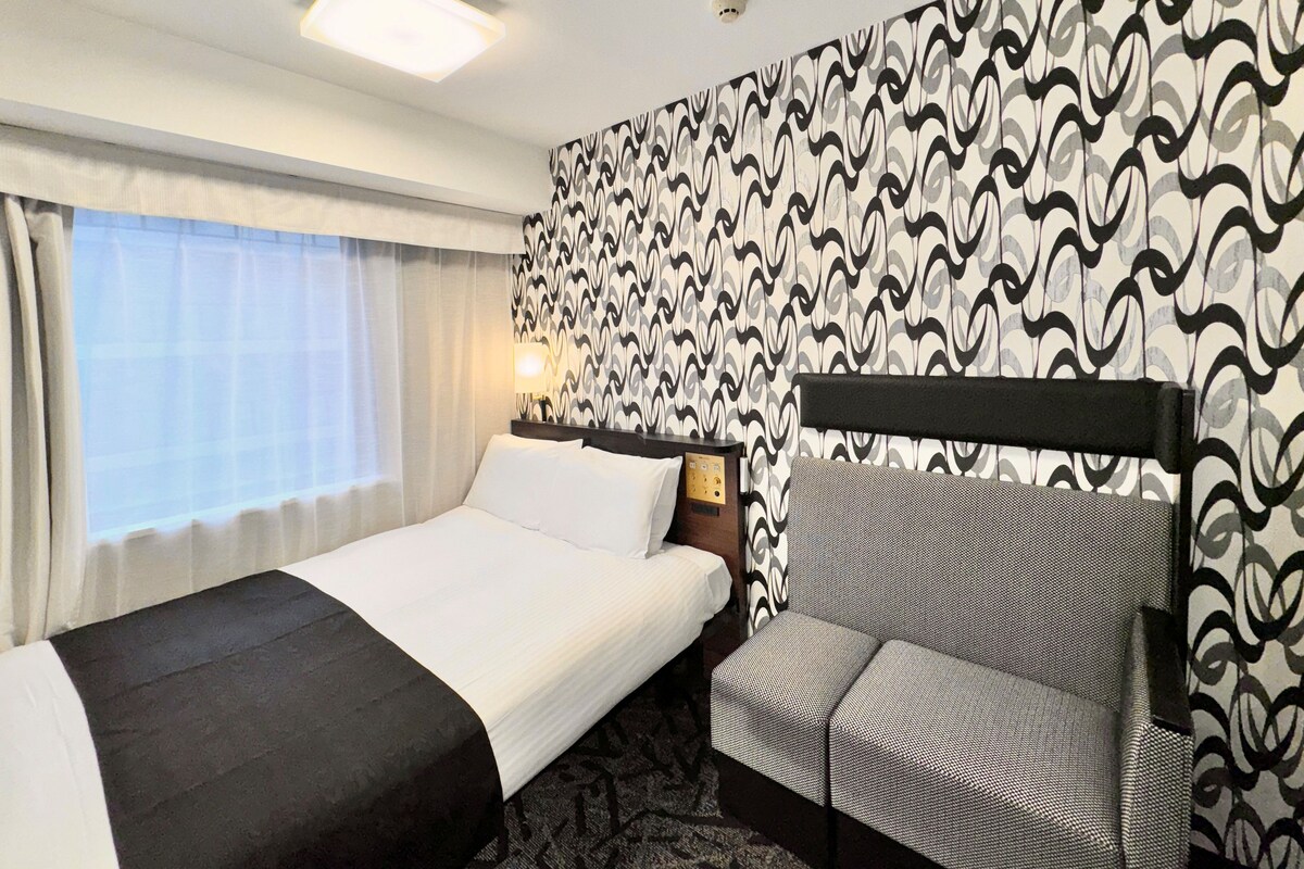 A hotel room with a bed, a chair, a bedside table with a lamp, and a patterned wall.