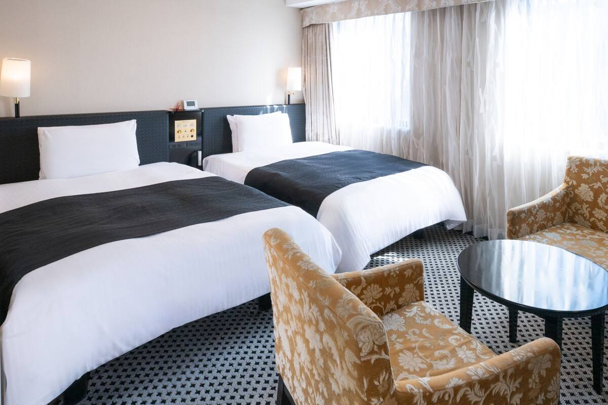 A hotel room with two beds, a small table, two chairs, a nightstand with a lamp, and a window with curtains.
