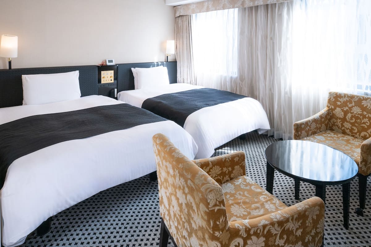 Quarto de hotel com duas camas, uma mesa pequena, duas cadeiras, uma mesinha de cabeceira com abajur e uma janela grande com cortinas.