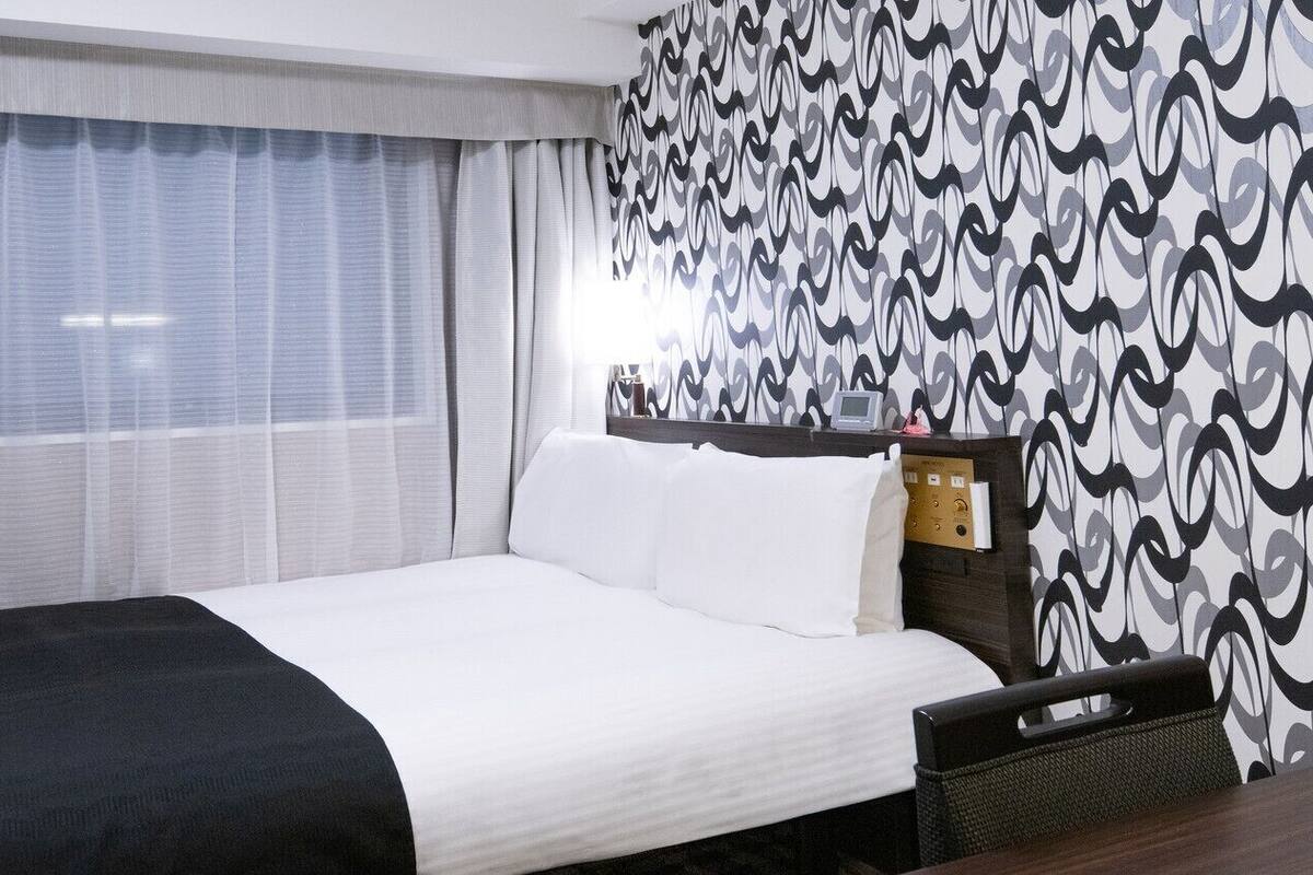 Une chambre d’hôtel avec un lit, une tête de lit dotée d’un panneau de commande, une fenêtre avec des rideaux légers et un mur à motifs.