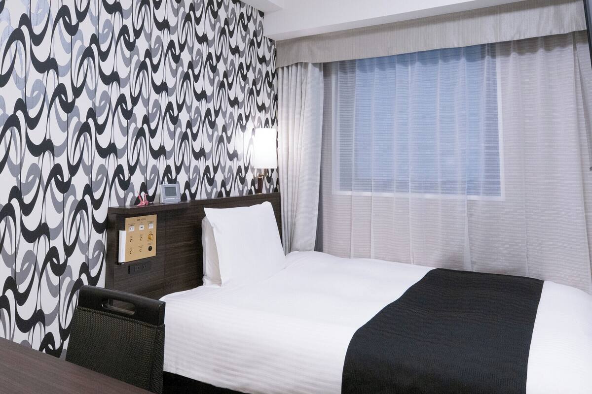 Une chambre d’hôtel comprenant un lit, une table de chevet, un bureau, une fenêtre avec des rideaux et un mur recouvert d’un papier peint à motifs.