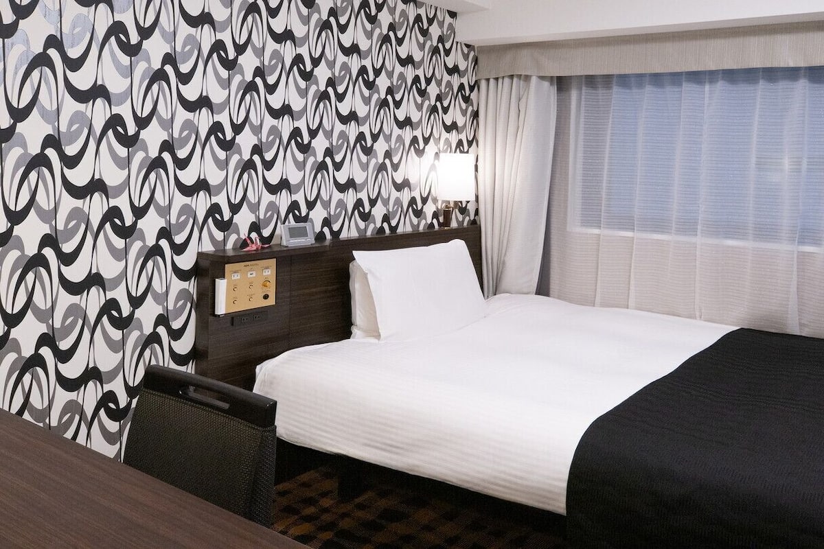 Une chambre d’hôtel avec un grand lit, un bureau, une chaise, un mur décoré de motifs et une fenêtre avec des rideaux.