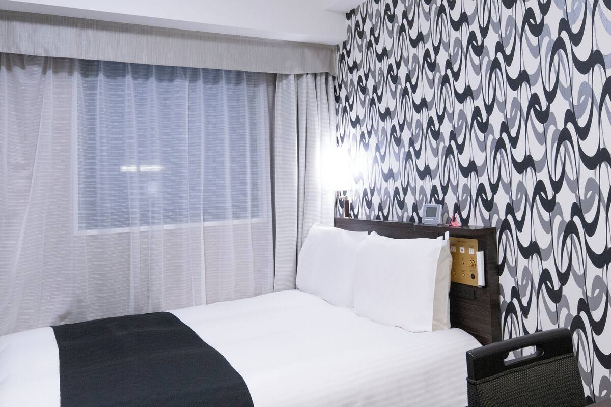 Une chambre d’hôtel avec un lit, un bureau, une chaise, une fenêtre avec des rideaux et un mur décoré d’un motif.