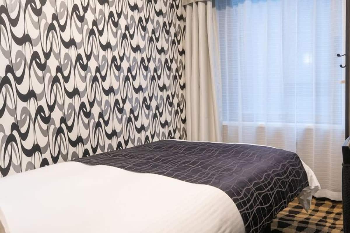 A hotel room with a patterned wall, a bed with a dark patterned bedspread, a chair, and a window with curtains.