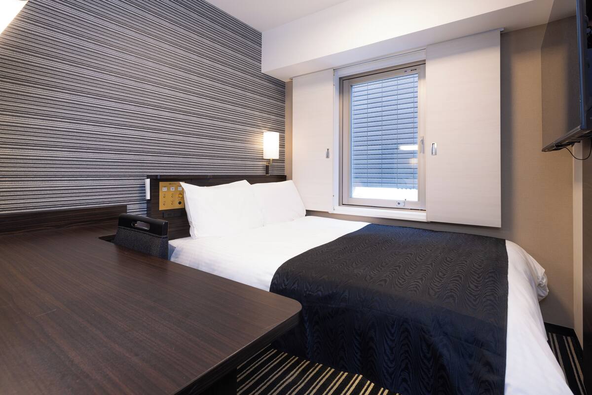 Hotellihuone, jossa on sänky, yöpöytä, lamppu ja ikkuna, jossa on sälekaihtimet.