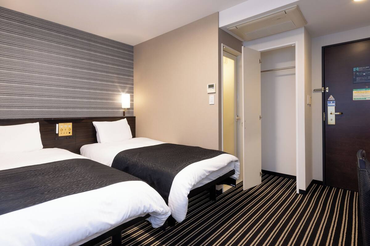 Hotellihuone, jossa on kaksi sänkyä, ohjauspaneeli, ovi ja raidallinen matto.