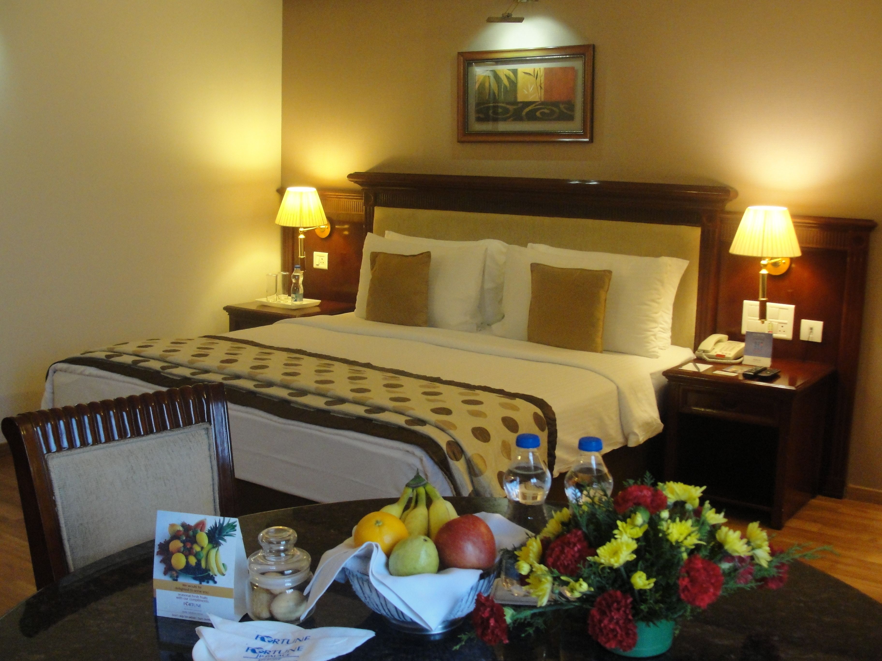 A hotel room with a bed, two bedside lamps, a nightstand with a phone, a table with a fruit bowl, a vase of flowers, and a framed picture on the wall.