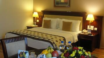 A hotel room with a bed, two bedside lamps, a nightstand with a phone, a table with a fruit bowl, a vase of flowers, and a framed picture on the wall.