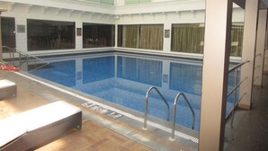 Ein Hallenbad mit klarem, blauen Wasser, umgeben von einer weiß gefliesten Fläche mit einem Rettungsschwimmerstuhl und einer Bank.