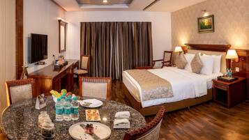 A hotel room with a bed, bedside tables, a desk, a chair, a dining table with water bottles and a dessert plate, and a large window with curtains.