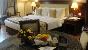 Ein Hotelzimmer mit einem ordentlich bezogenem Bett, zwei Nachttischen mit Lampen, einem kleinen runden Tisch mit Obst und einer Blumenvase, sowie einem gerahmten Bild an der Wand.