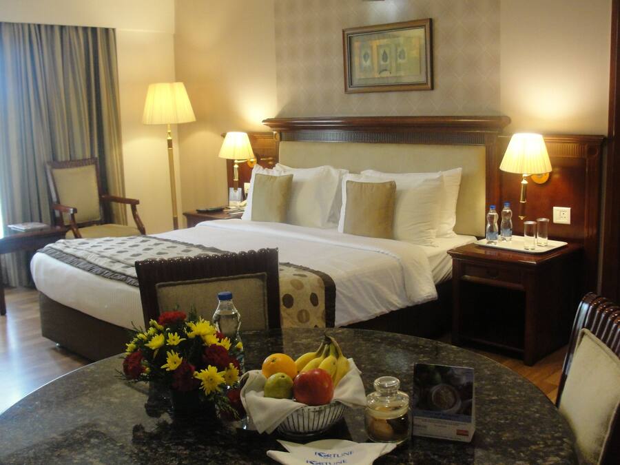 A hotel room with a neatly made bed, two bedside tables with lamps, a small round table with fruit and a vase of flowers, and a framed picture on the wall.