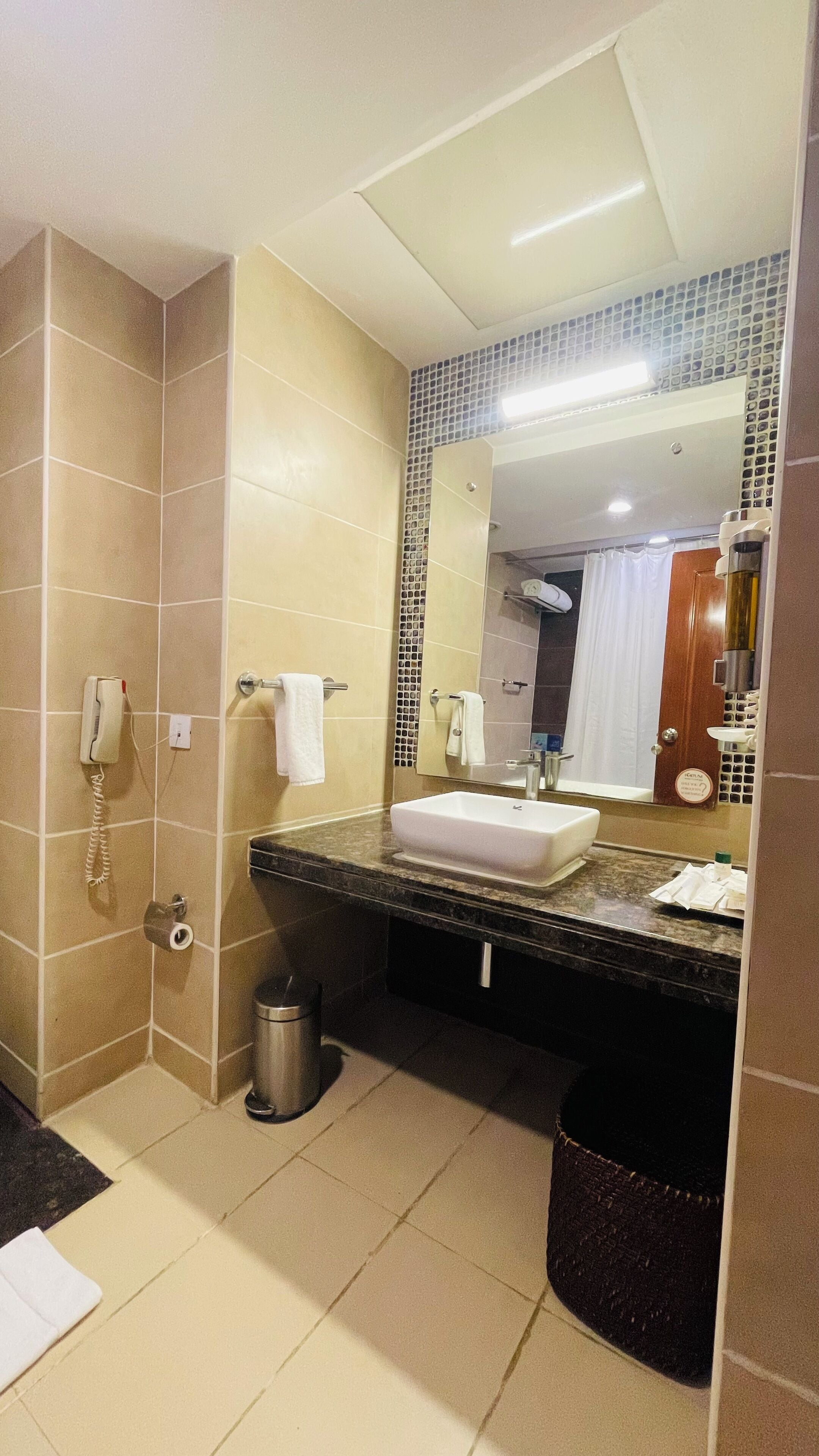 Un baño con lavamanos blanco, un espejo, un teléfono y un cesto de basura.