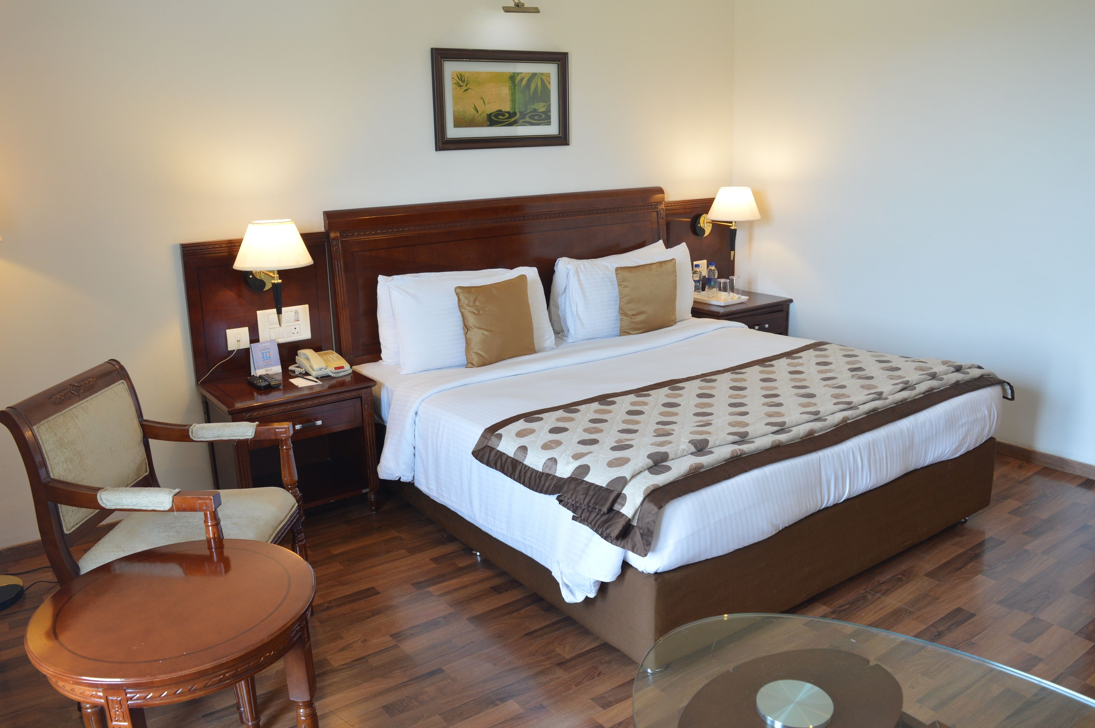 A hotel room with a large bed, two bedside tables with lamps, a wooden chair, and a glass table.