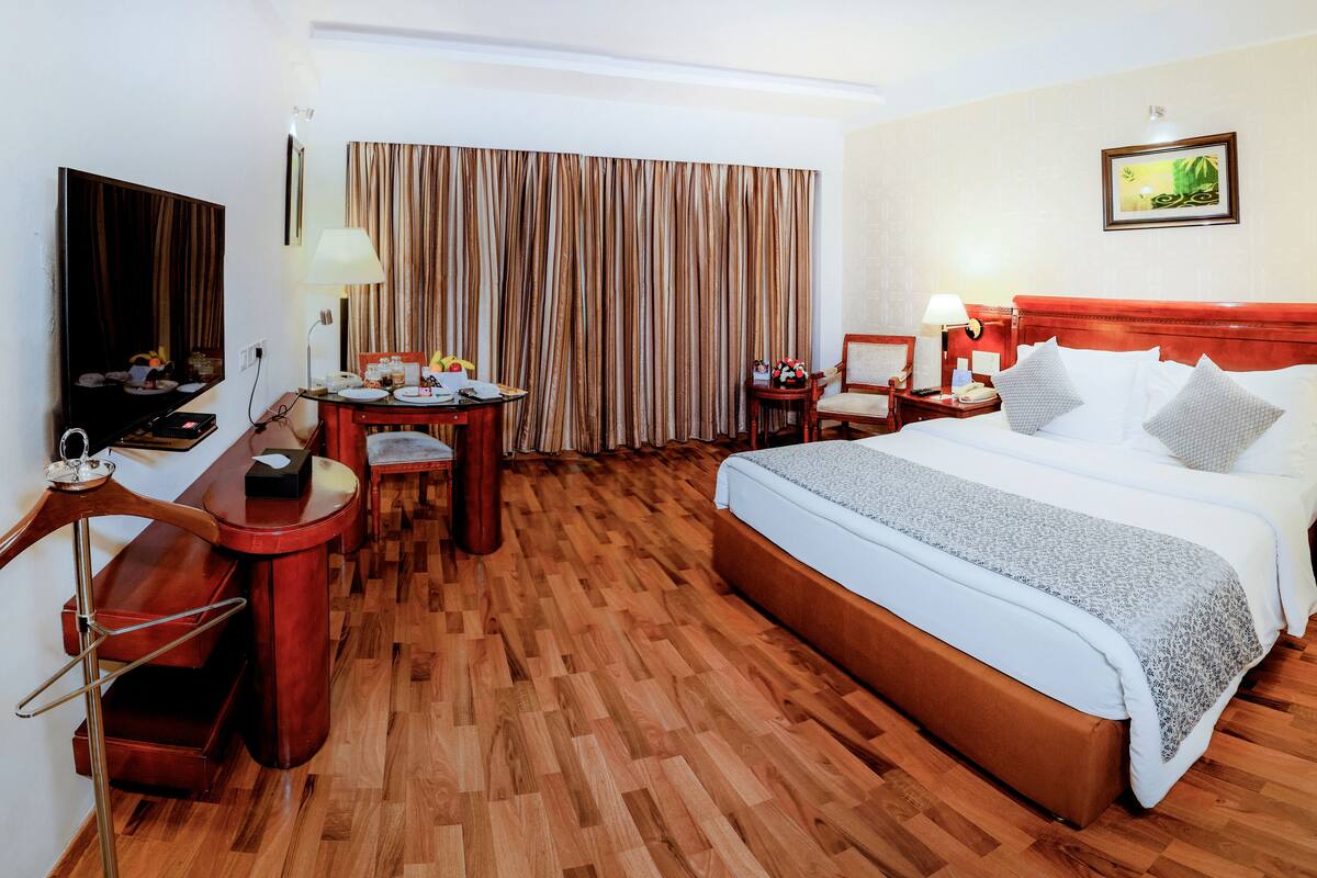 Une chambre d’hôtel avec un plancher en bois, un lit avec une literie blanche, un bureau avec une chaise, une petite table avec une lampe et une grande fenêtre avec des rideaux.