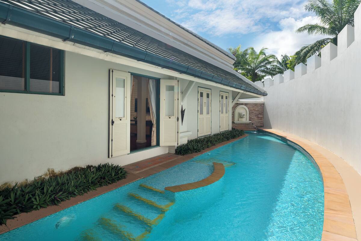 Piscina junto a un edificio con varias puertas y ventanas, rodeada por un muro blanco y vegetación.