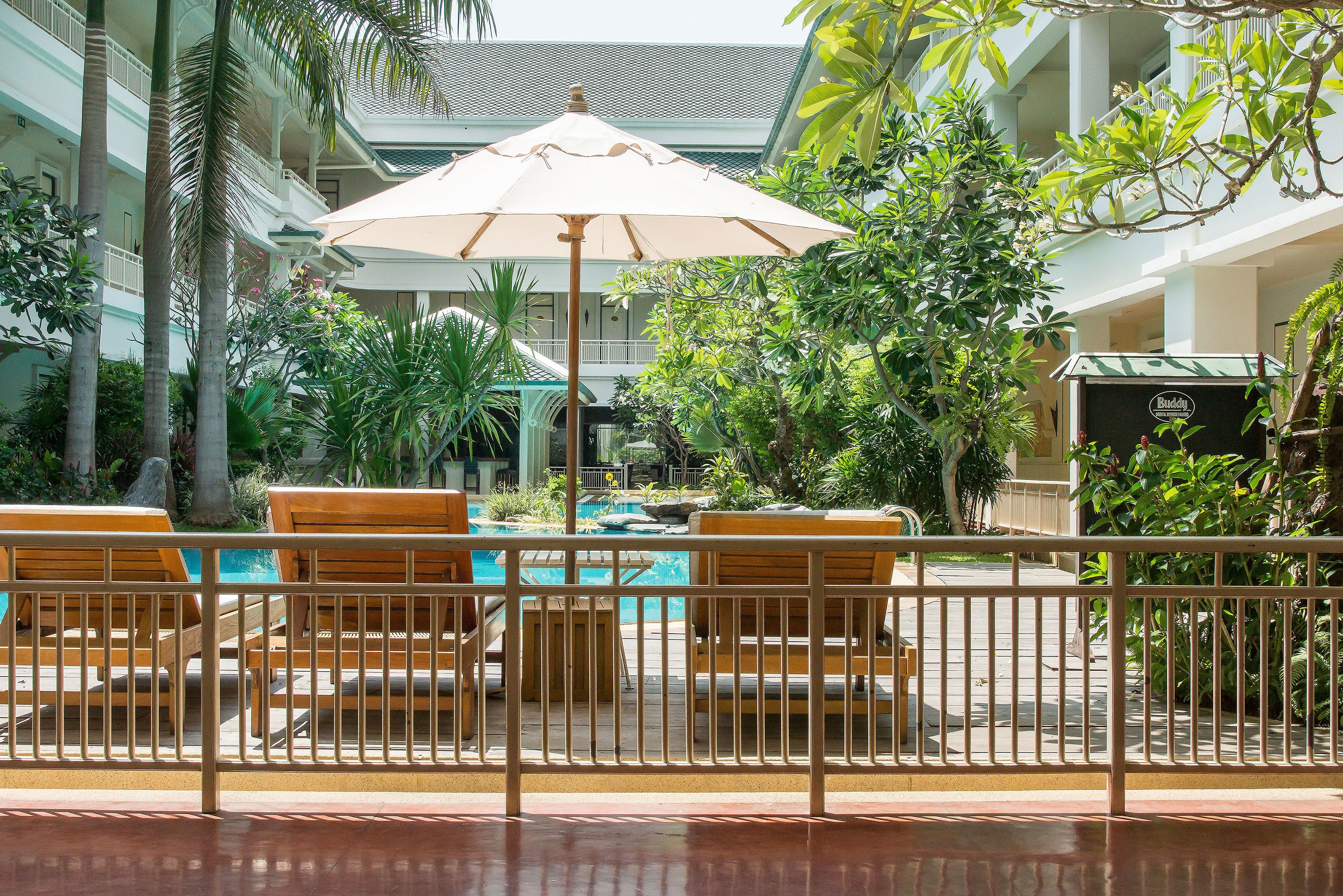 A pool area with wooden chairs and an umbrella, surrounded by lush greenery and a white railing.