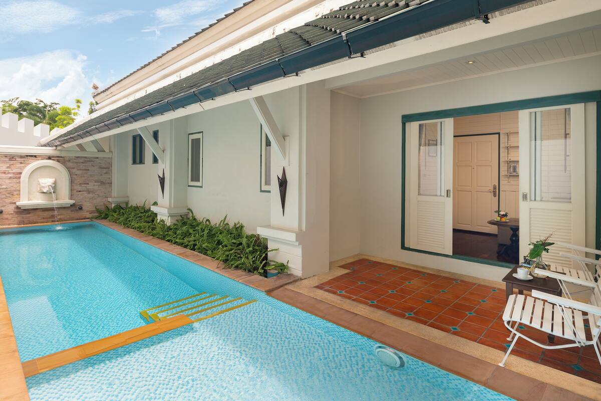Una piscina con un borde de baldosas, una pequeña mesa con una planta y una puerta que da a una habitación.