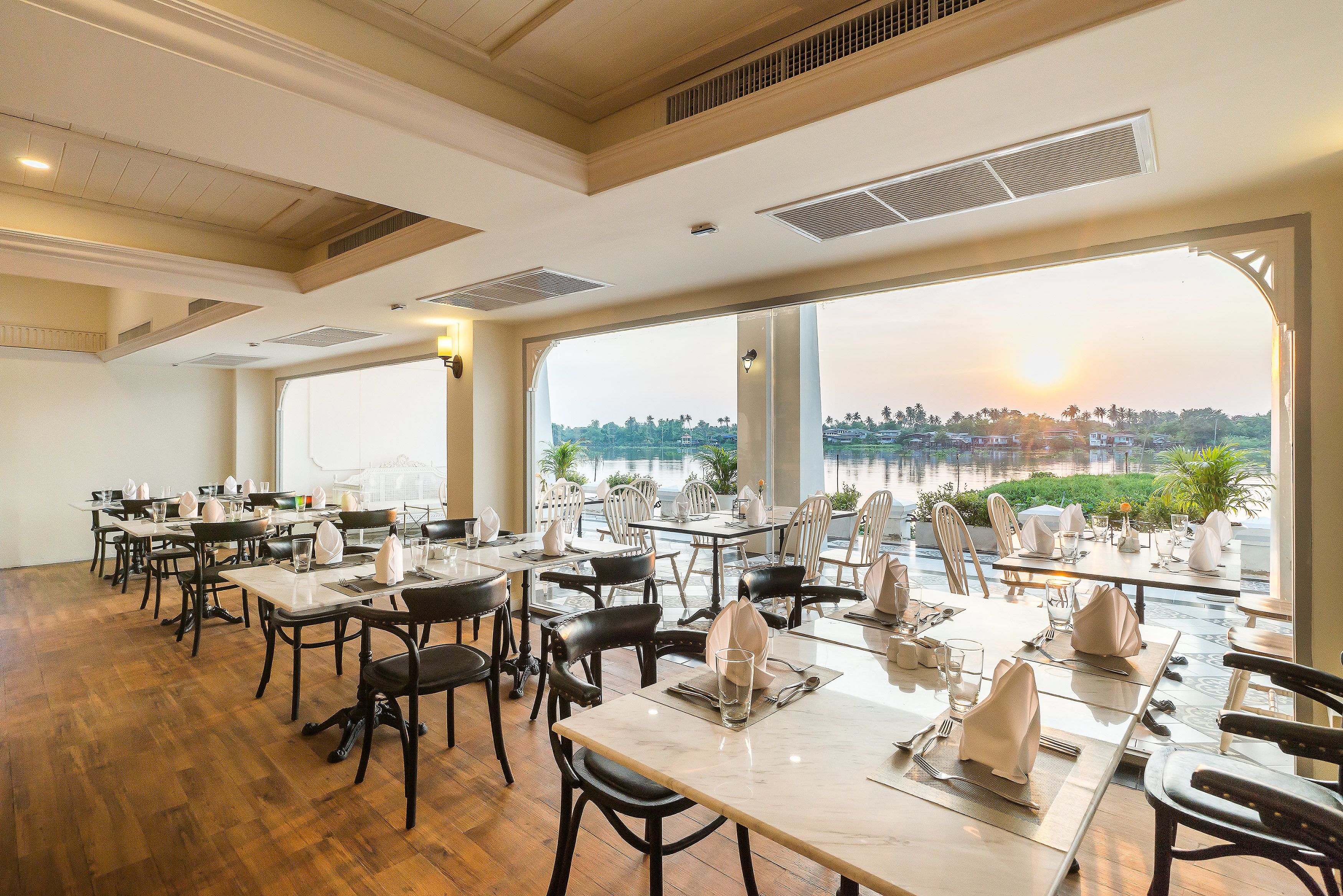 A restaurant with tables set for dining, large windows with a view of a body of water and greenery, and a wooden floor.