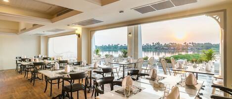 A restaurant with tables set for dining, large windows with a view of a body of water and greenery, and a wooden floor.