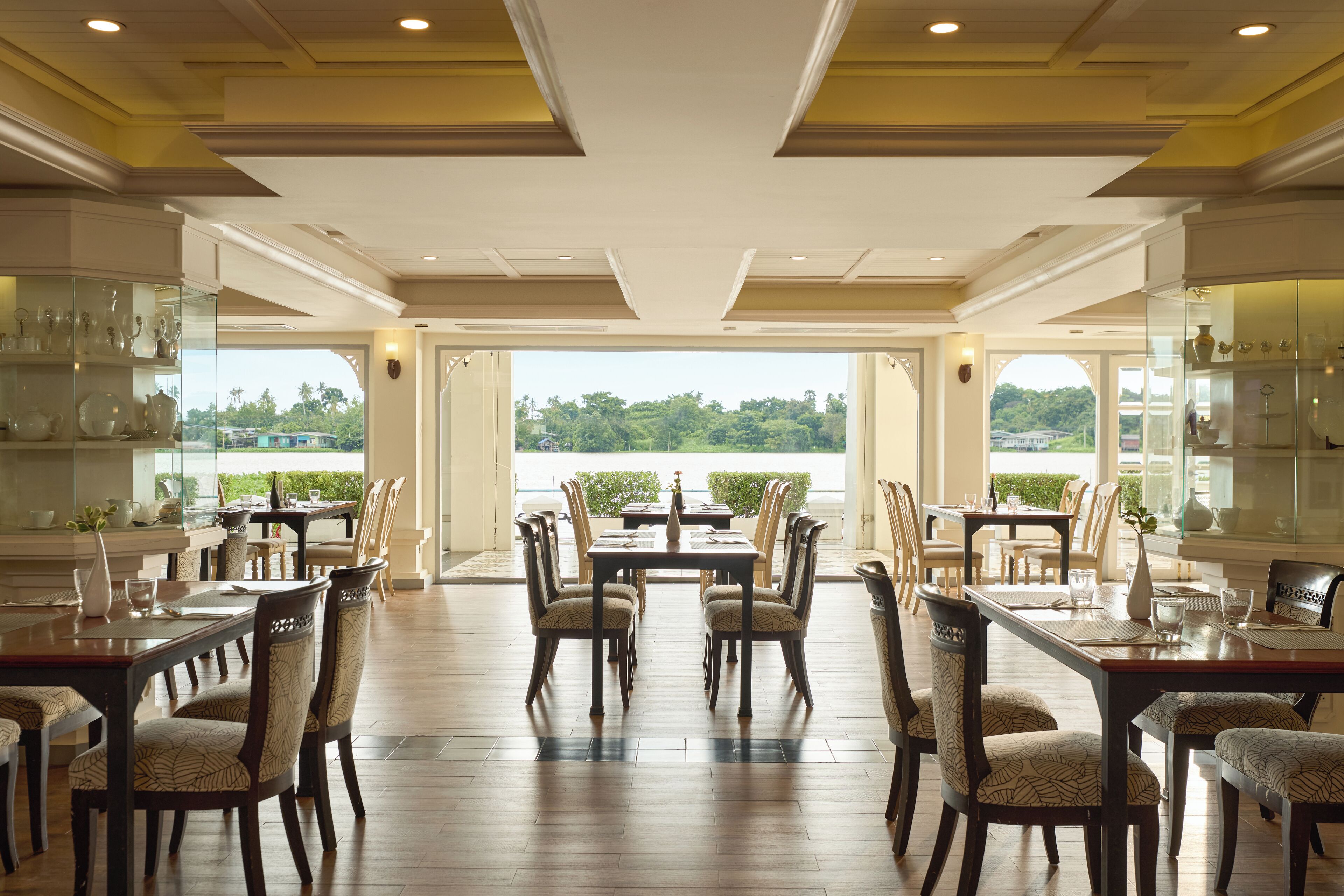 A spacious dining area with multiple tables and chairs, large windows offering a view of greenery and a body of water.