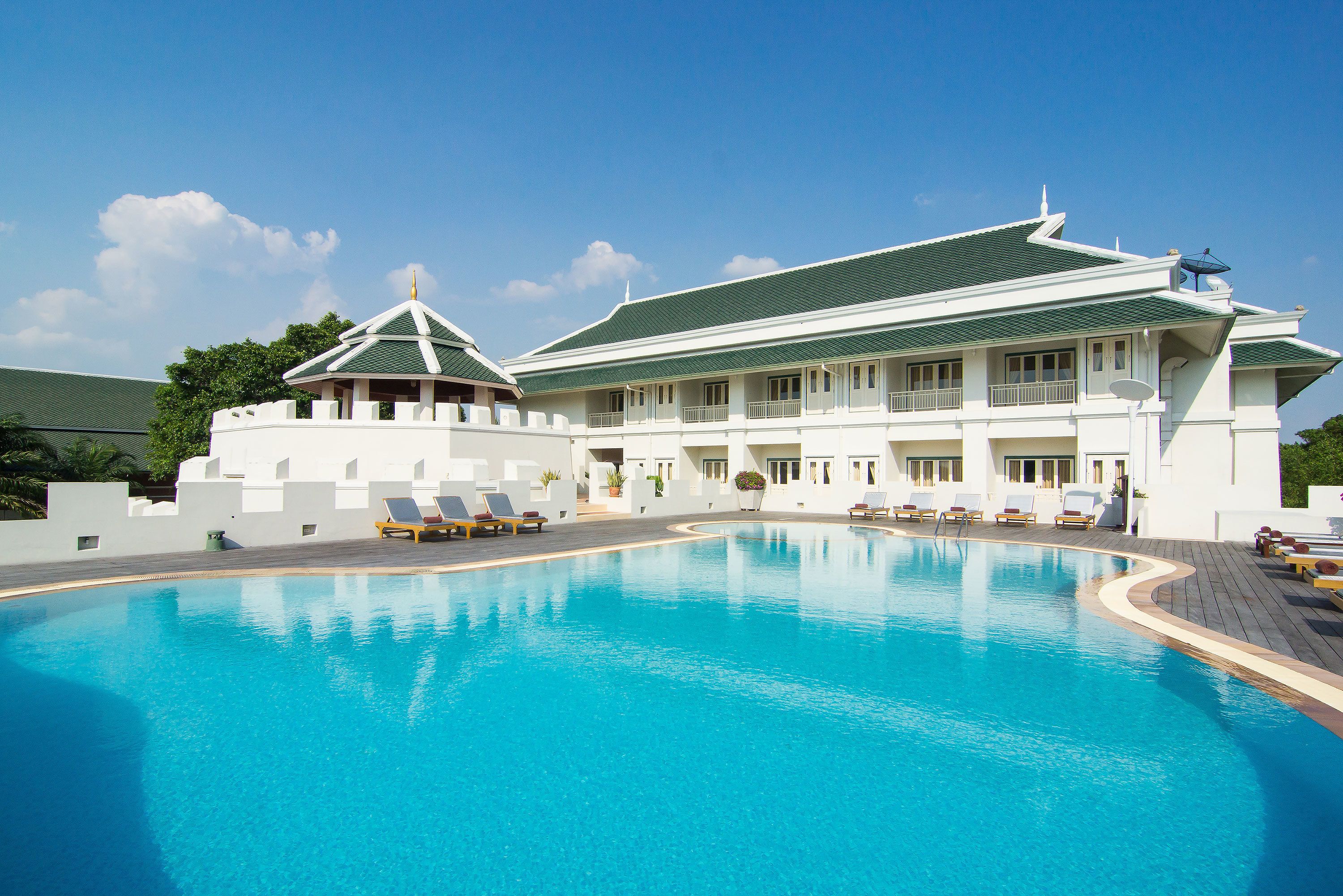 A large swimming pool with lounge chairs and a building with a green roof and white walls.