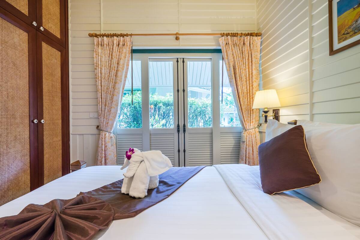 Una habitación de hotel con cama, ventana con cortinas, lámpara y un cuadro en la pared.