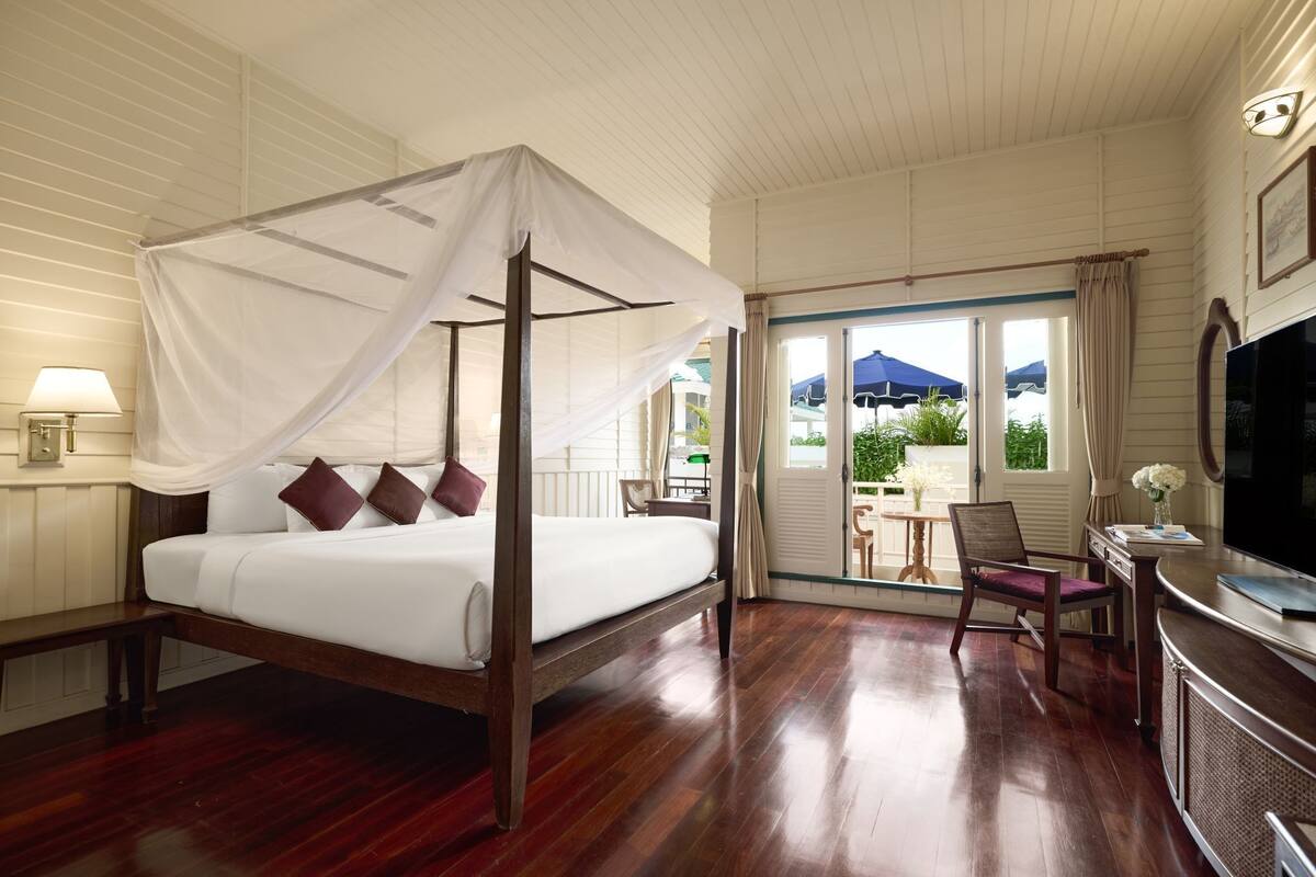 Una cama con dosel, televisor, escritorio y silla en una habitación de hotel con piso de madera y un balcón con vistas a follaje.