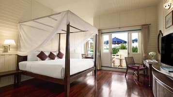 A four-poster bed with a canopy, a television, a desk, and a chair in a hotel room with wooden flooring and a balcony with a view of greenery.