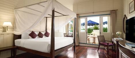 A four-poster bed with a canopy, a television, a desk, and a chair in a hotel room with wooden flooring and a balcony with a view of greenery.