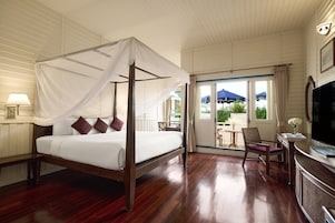A four-poster bed with a canopy, a television, a desk, and a chair in a hotel room with wooden flooring and a balcony with a view of greenery.