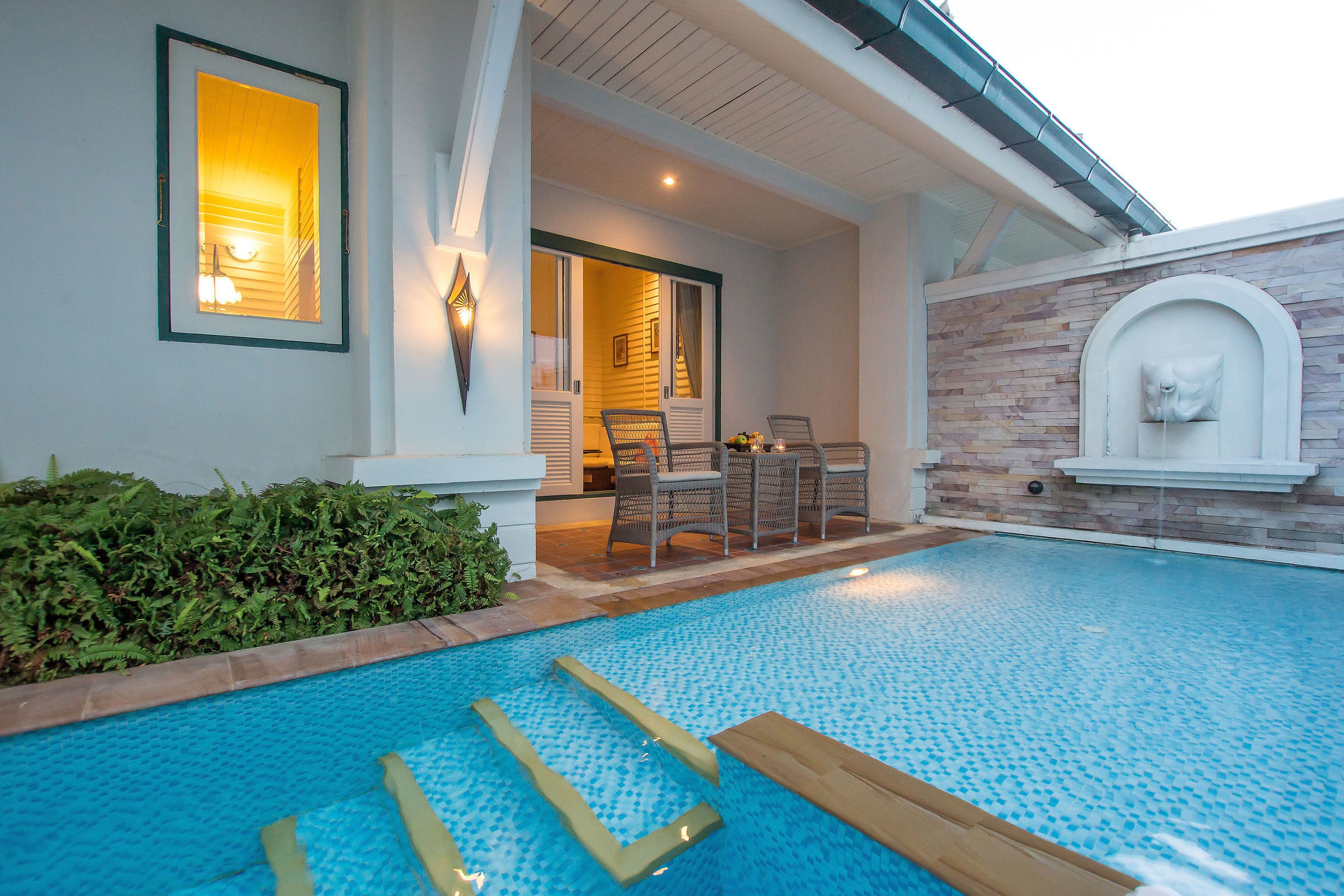A swimming pool with steps leading into it, a covered patio area with outdoor furniture, and a stone wall with a mailbox.