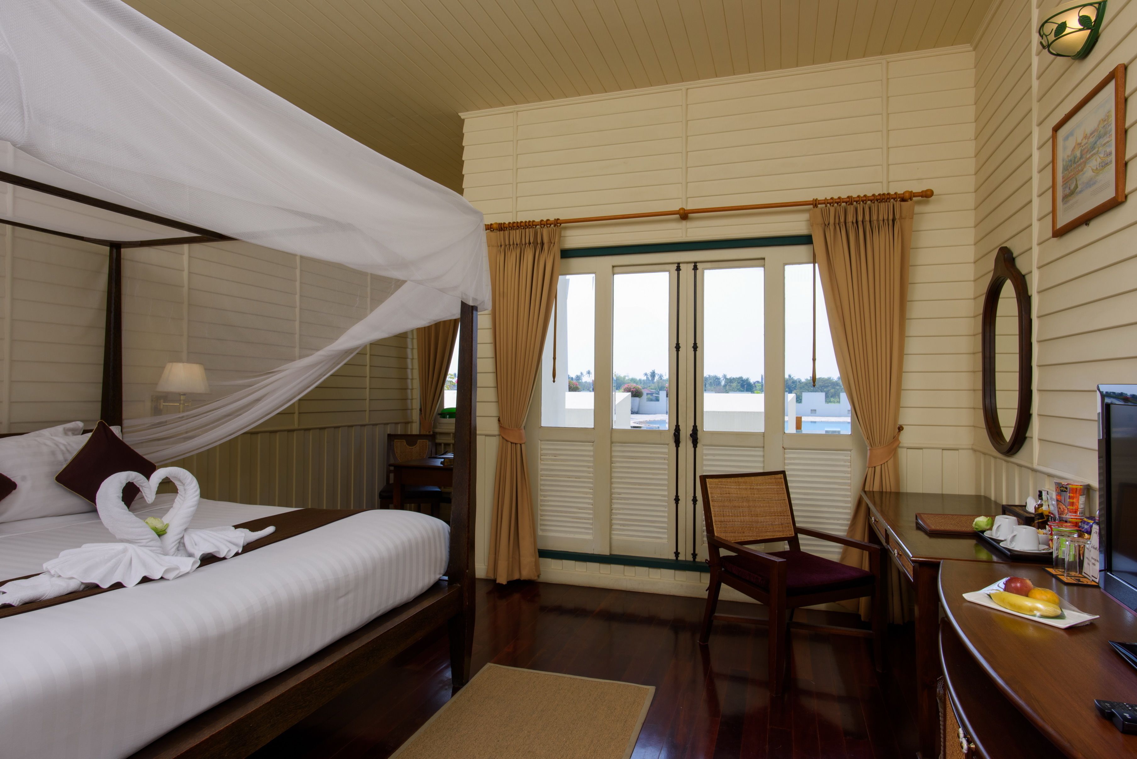Une chambre d’hôtel avec un lit à baldaquin, un bureau avec une chaise et une vue sur l’extérieur, visible à travers une porte munie de rideaux.