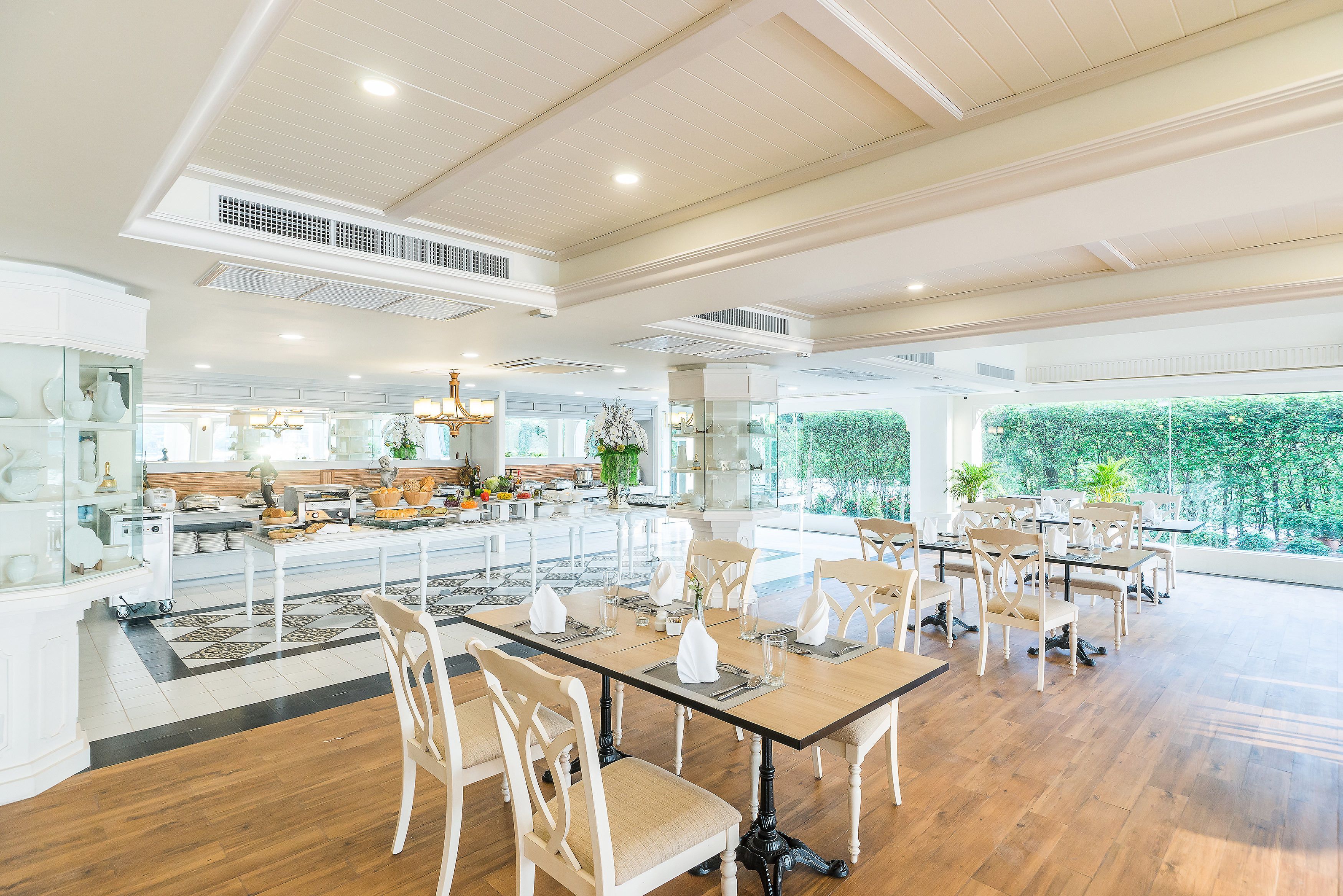 A spacious dining area with multiple tables and chairs, a kitchen in the background, and large windows offering a view of greenery.
