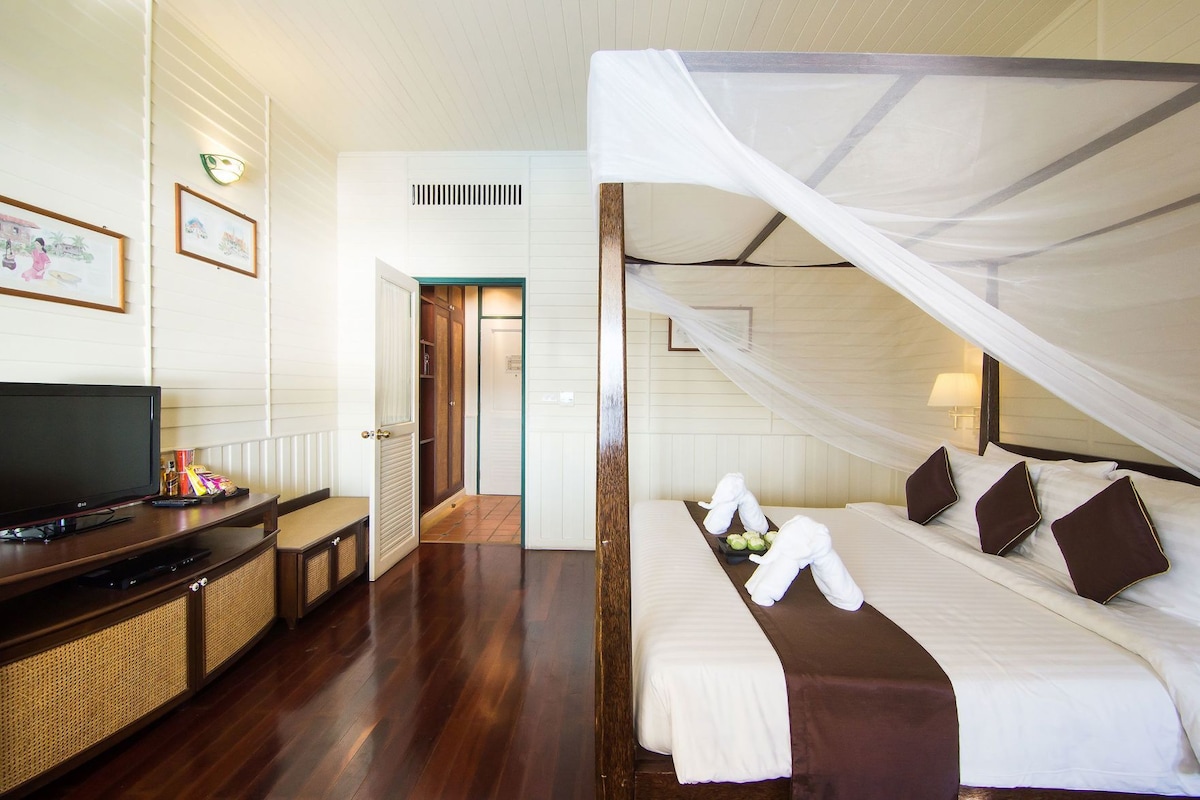 Quarto de hotel com televisão, cama com roupa de cama branca e castanha, mesa de cabeceira e cama com dossel.