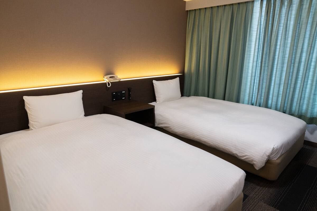 Une chambre d’hôtel avec deux lits simples, une table de chevet avec un téléphone et une fenêtre avec des rideaux.