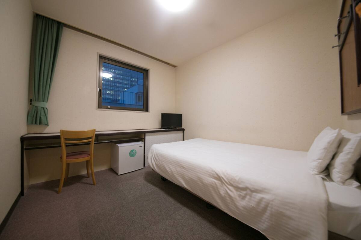 Een kleine, schone kamer met een enkel bed, een bureau, een stoel en een kleine koelkast.