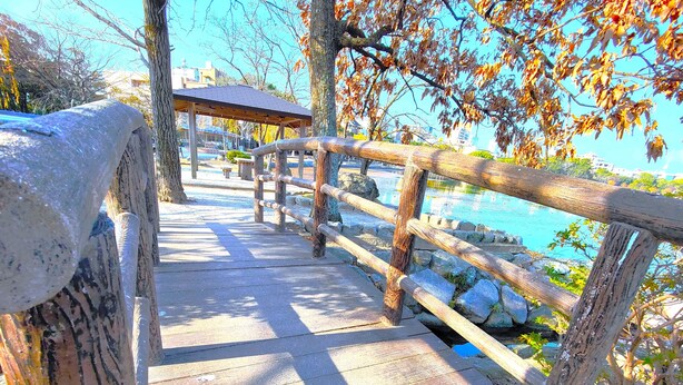 Eine Holzbrücke mit Steinbrüstung, die zu einem hölzernen Pavillon an einem Gewässer führt.