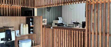 A modern reception area with a wooden slat partition, a digital kiosk, a coffee machine, and a counter with a cash register.