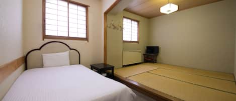 A room with a bed, a small table, a futon, a TV, and a window with a view of the outside.