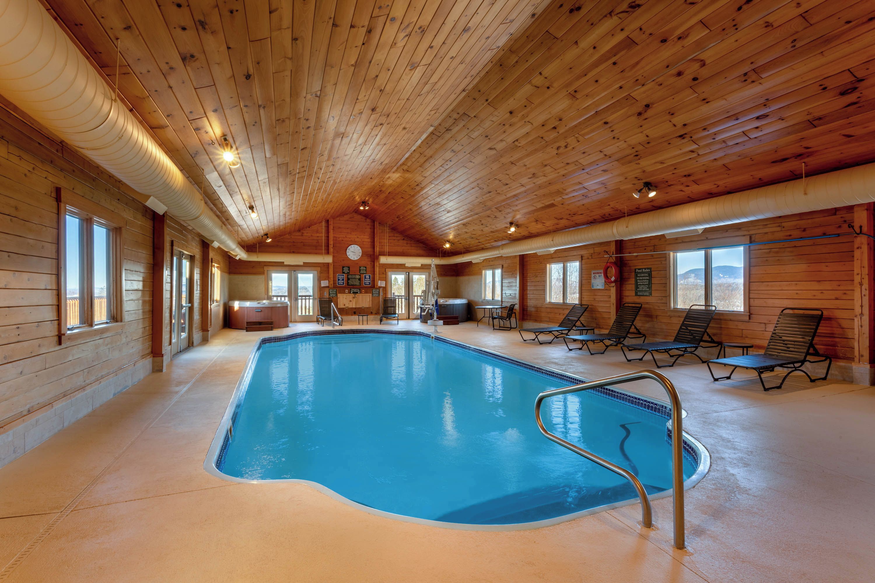 Une piscine intérieure avec un revêtement intérieur en bois, une horloge au mur et des chaises longues disposées autour de la piscine.