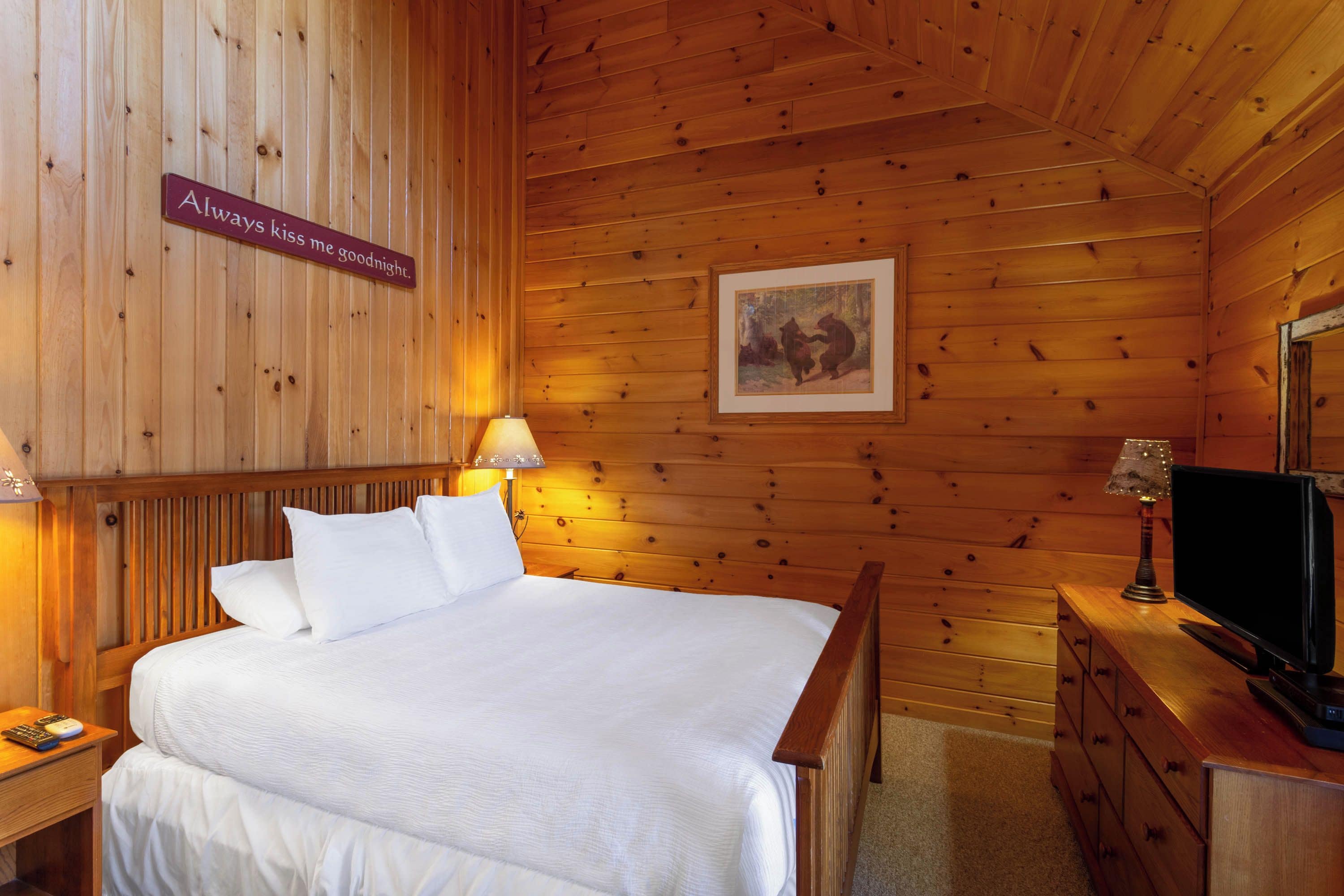 A wooden room with a bed, a nightstand, a lamp, a dresser, and a television.