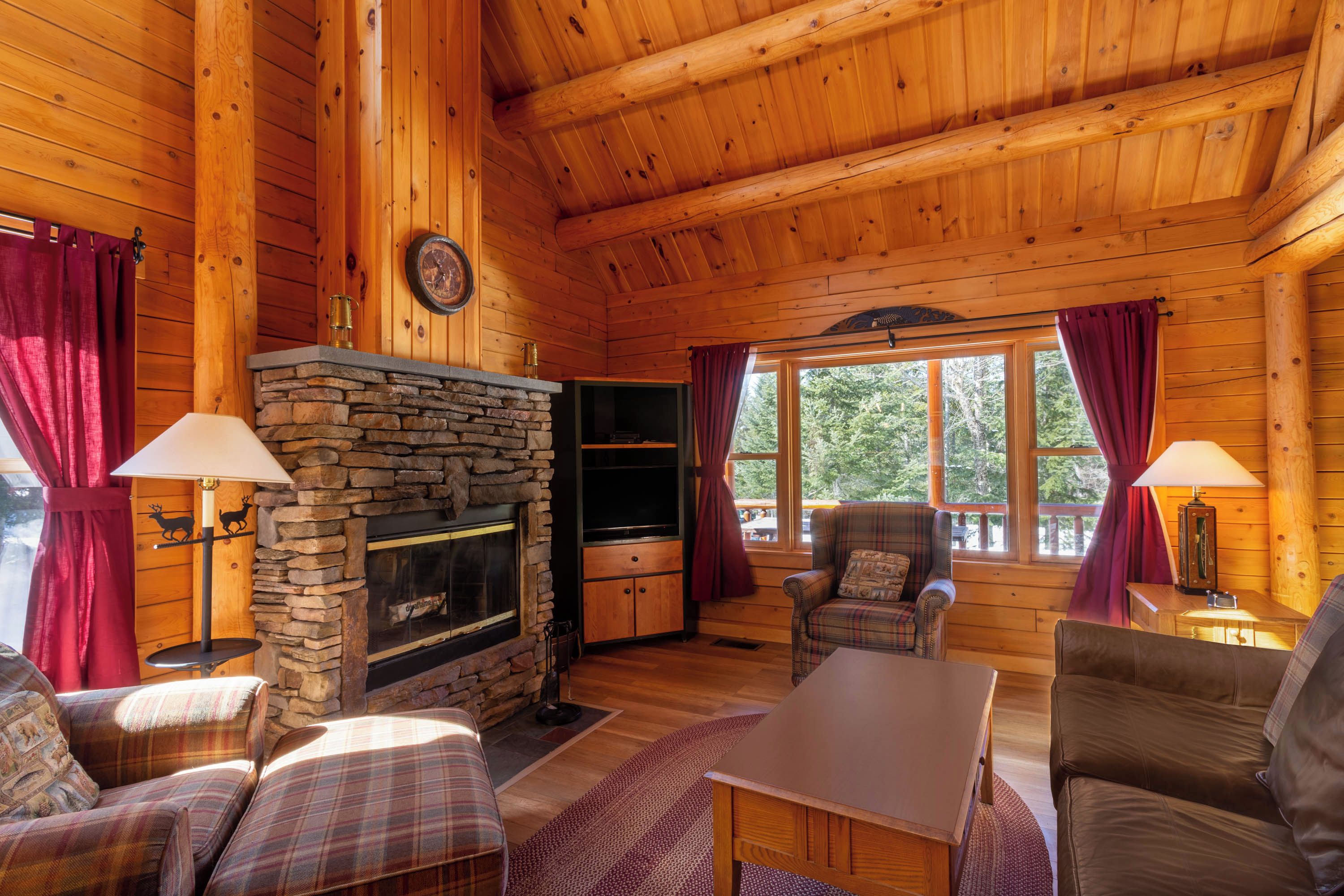 A cozy cabin interior with a stone fireplace, plaid furniture, and large windows.