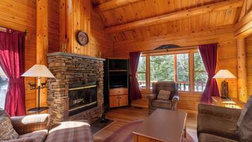 A cozy cabin interior with a stone fireplace, plaid furniture, and large windows.