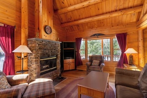 A cozy cabin interior with a stone fireplace, plaid furniture, and large windows. - Rangeley Lake Resort (Rangeley)