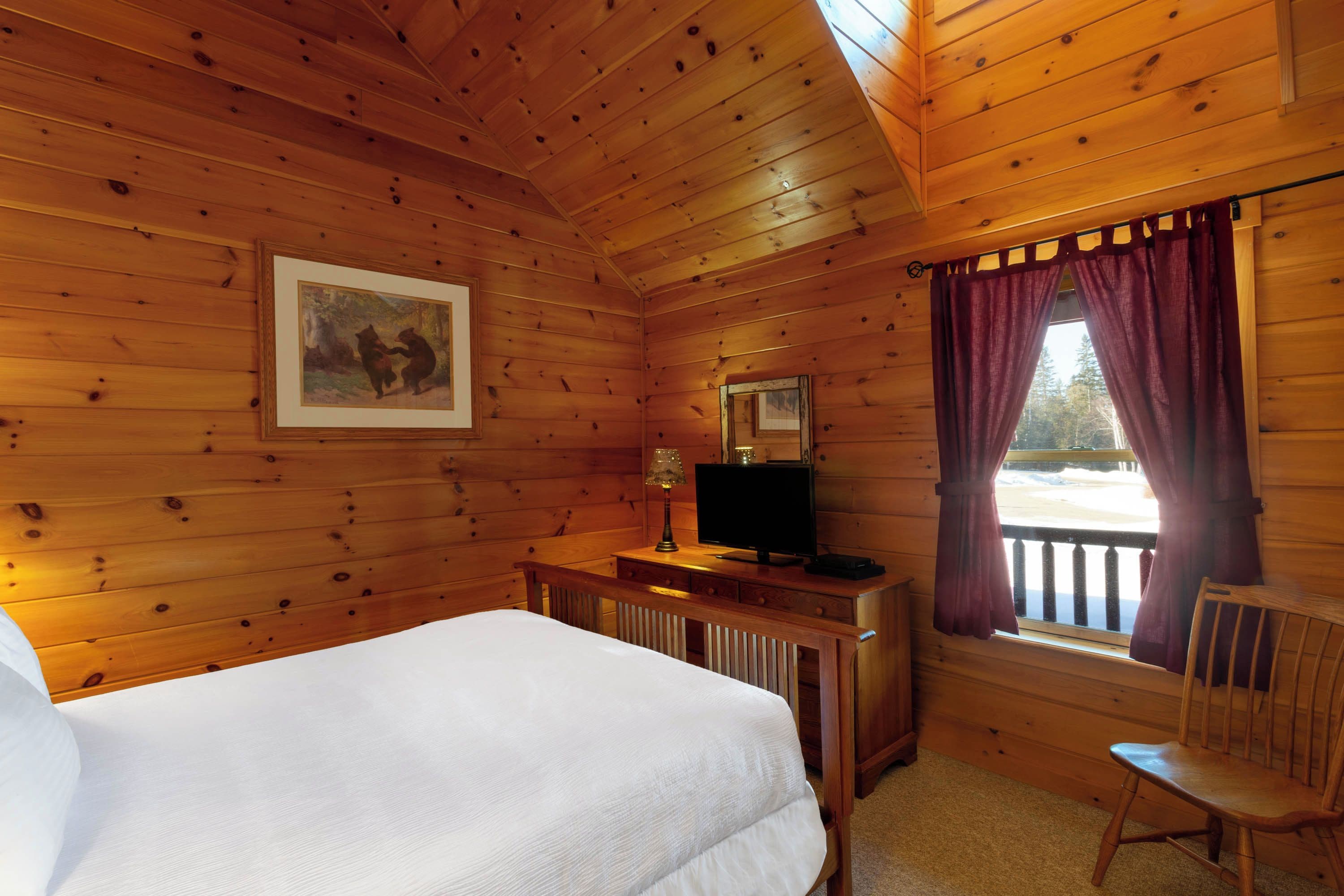 A wooden cabin room with a bed, a desk with a television, a chair, and a window with curtains.