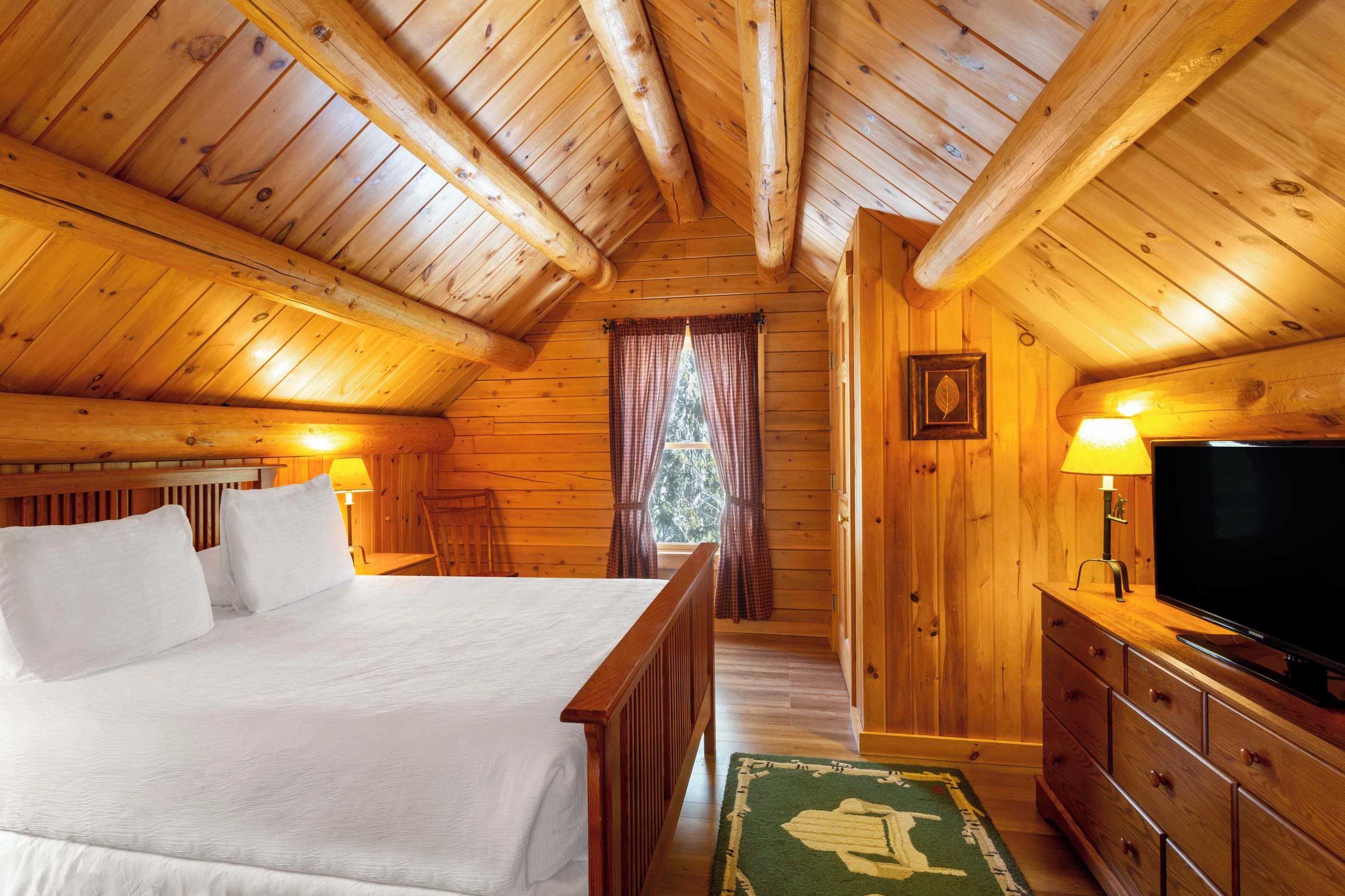 Habitación de cabaña de madera con cama, televisor y ventana con cortinas.