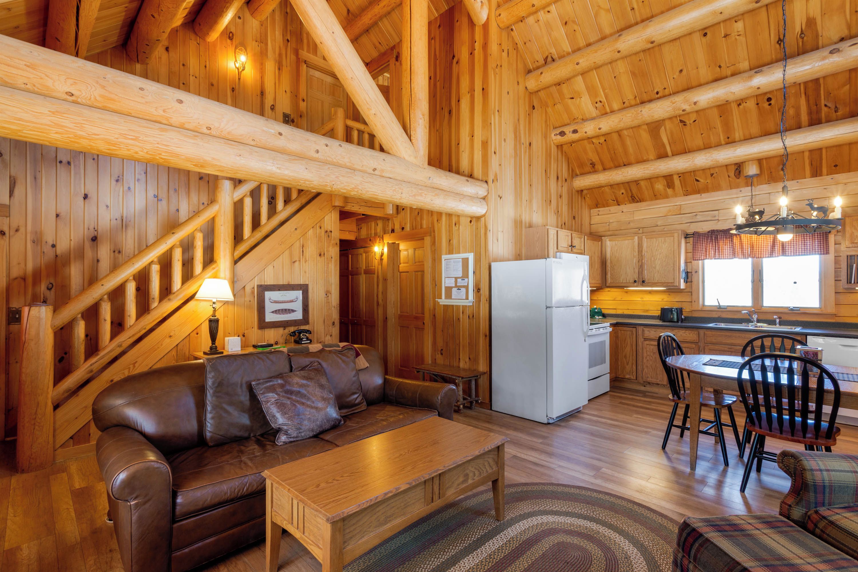 A cozy cabin interior with a wooden staircase, a dining area, a kitchen with a refrigerator, and a living room with a leather sofa and a coffee table.