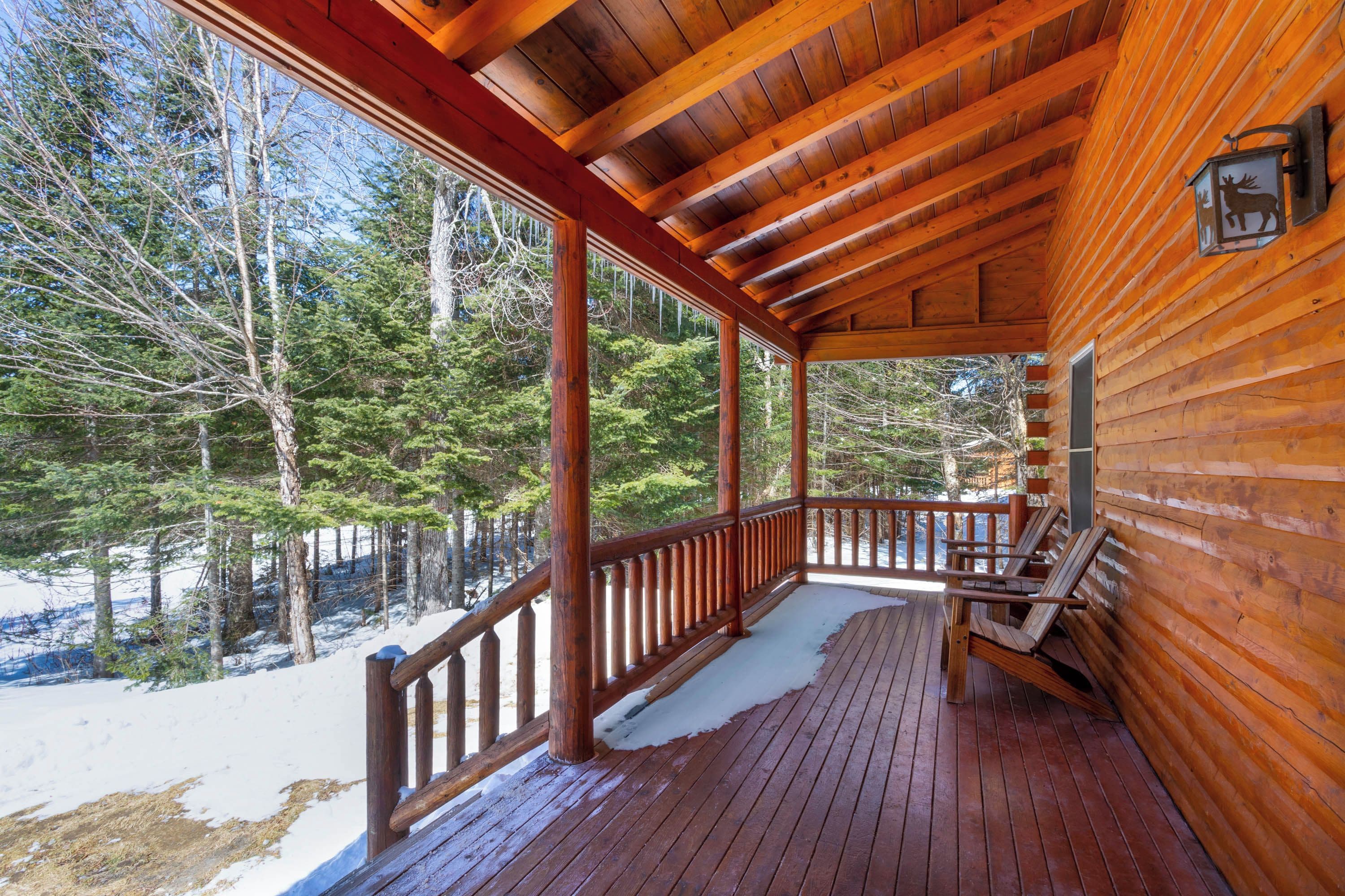Une cabane en bois dotée d’un porche couvert, d’un banc et offrant une vue sur une forêt enneigée.